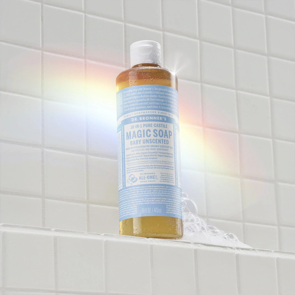 A bottle of Dr. Bronners Baby Unscented Castile Soap is placed upright on a white tiled shower shelf, with a faint rainbow reflection appearing nearby [16 oz]