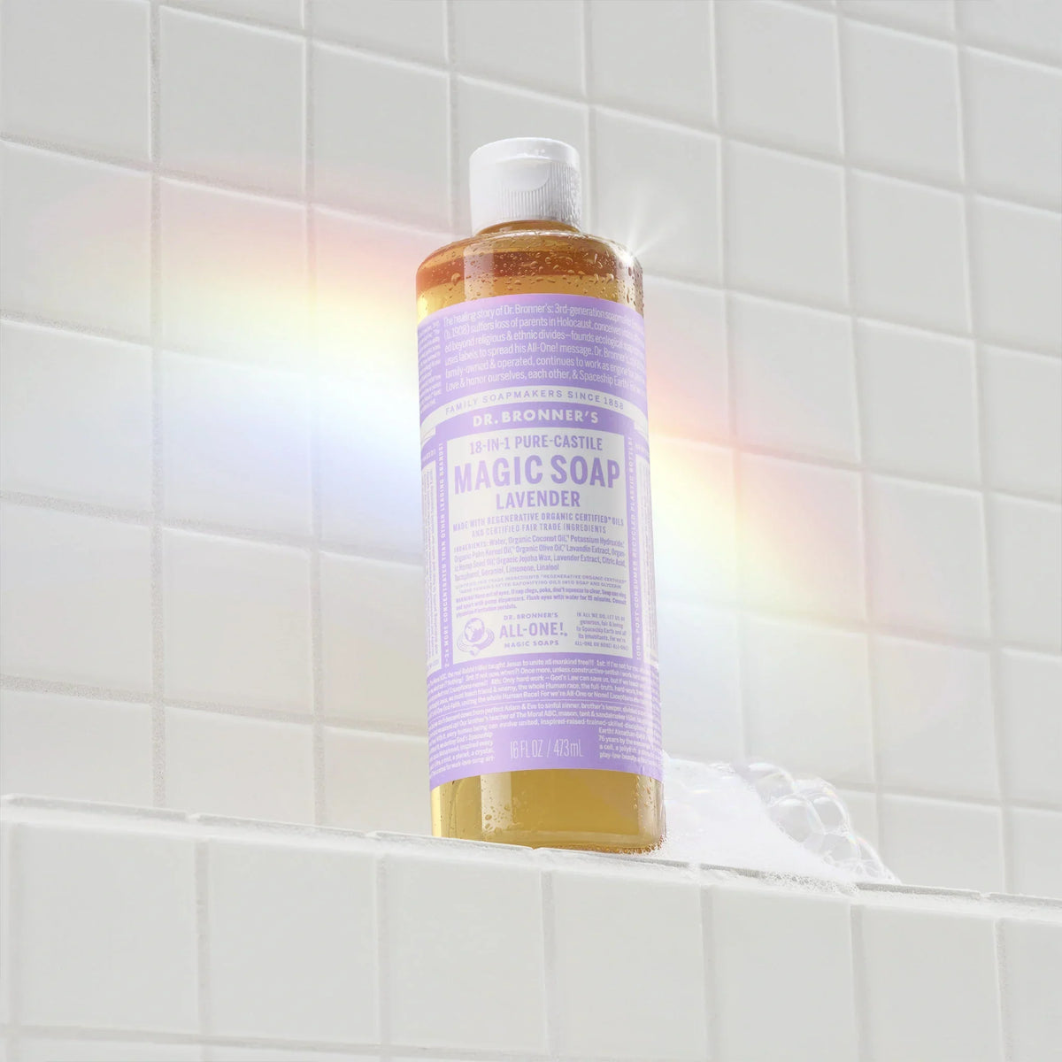 A bottle of Dr. Bronners lavender liquid castile soap sits on a tiled shower shelf, illuminated by a soft rainbow light. The label features text highlighting its organic, eco friendly ingredients [16 oz]