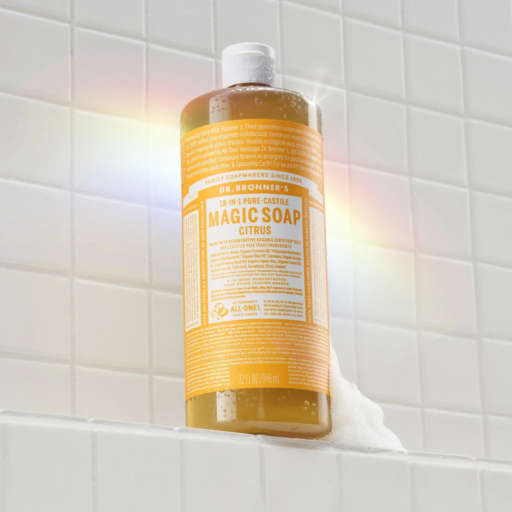 A bottle of Dr. Bronners 18 in 1 Pure Castile Magic Soap Citrus stands on a tiled surface with soap bubbles forming around its base, illuminated by soft rainbow light [32 oz]