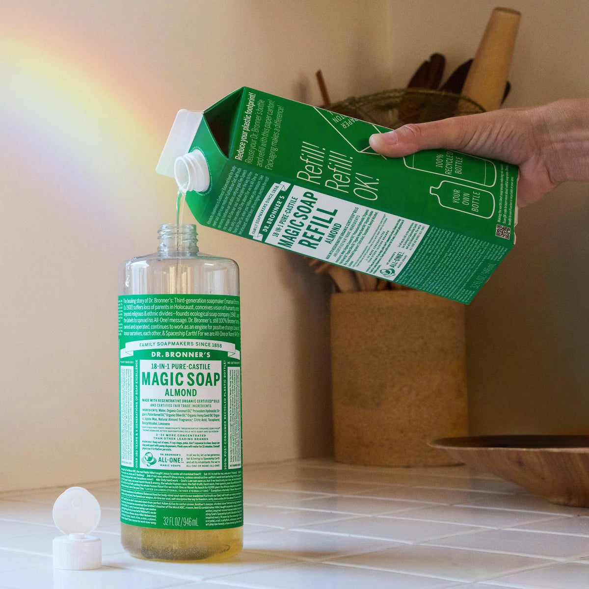 A hand pours Dr. Bronners Almond Pure Castile Liquid Soap into a bottle from a refill container, set on a white tiled surface with kitchen utensils in the background. Text includes MAGIC SOAP, ALMOND, Refill Refill Refill Ok and ALL ONE Keywords Dr. Bronners almond castile soap, liquid castile soap, biodegradable soap [32 oz]